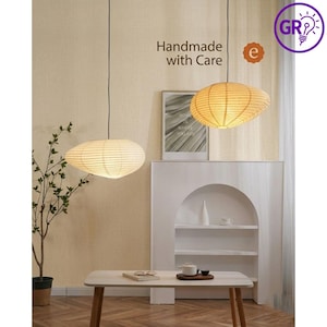 Puede incluir: Dos lámparas colgantes con diseño de farol de papel cuelgan del techo. Las lámparas ovaladas emiten un brillo cálido. El texto "Handmade with Care" es visible. Las luces están suspendidas sobre una mesa blanca.
