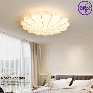 May include: A white, petal-shaped ceiling light fixture with a soft, diffused glow. The light is mounted on a white ceiling in a bedroom setting. The text "MODERN DESIGN" and "MANIFOLD BLESSINGS" is visible.