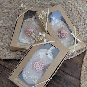 Peut inclure: Trois désodorisants décoratifs en forme d'étiquette dans des boîtes en carton. Chaque désodorisant est rose clair avec un motif floral et des accents blancs. Les boîtes sont attachées avec de la ficelle et ornées de fleurs séchées. Le texte "Suspension parfumée" est visible.