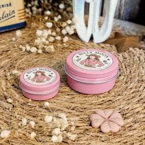 Può includere: Sono esposti due barattoli di metallo rosa con coperchi, uno piccolo e uno grande. Ogni barattolo presenta un'etichetta con la scritta "BILLY MOON" e un'illustrazione di una persona. È visibile anche un oggetto rosa a forma di fiore. Gli oggetti sono su una superficie tessuta.