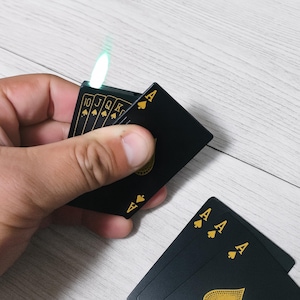 Op de afbeelding: Zwarte aansteker met speelkaarten thema en een groene vlam. De aansteker wordt in een hand gehouden, met de Aas van Schoppen kaart zichtbaar. Andere kaarten met gouden details zijn ook aanwezig, waaronder een reeks kaarten van 10 tot Koning.