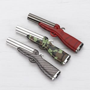 Op de afbeelding: Drie novelty aanstekers in de vorm van jachtgeweren. Eén is rood, één is camouflage en de derde heeft een koolstofvezelpatroon. Elk heeft een zilveren loop en trekker.