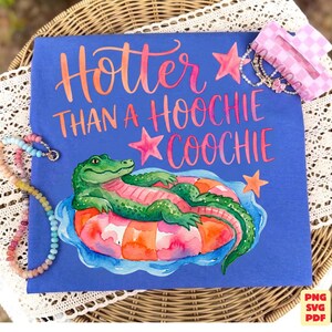 May include: A blue t-shirt with the slogan "Hotter Than A Hoochie Coochie" in orange and pink script. The shirt features a watercolour illustration of a green alligator on a pink and orange inflatable ring. A beaded necklace and a hair clip are also visible.