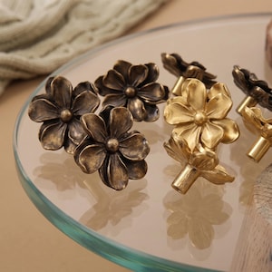 May include: A collection of metal flower-shaped cabinet knobs featuring a multi-layered petal design. The set includes pieces with a dark, antiqued bronze finish and others with a bright, polished gold-tone finish, each with a central stem.
