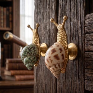 Peut inclure: Deux boutons de placard en forme d'escargot avec des corps en métal doré texturé et des coquilles en spirale naturelles. Une coquille est tachetée de vert et blanc, l'autre est crème avec des taches brunes. Chaque bouton a une base métallique.
