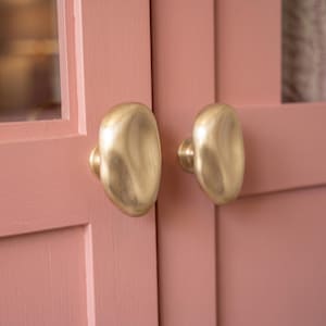 May include: Two oval-shaped cabinet knobs with a brushed gold finish attached to a pair of pink cabinet doors. The knobs have a smooth, organic, and slightly irregular surface texture, contrasting with the flat, matte pink painted wood surface.