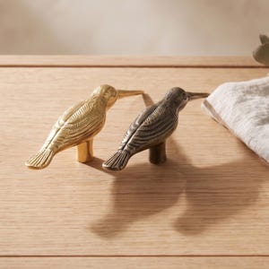 May include: Two bird-shaped cabinet knobs, one with a polished gold-tone finish and the other with a dark, antiqued bronze-tone finish. Each knob features detailed wing and feather textures, a long pointed beak, and a central cylindrical base.