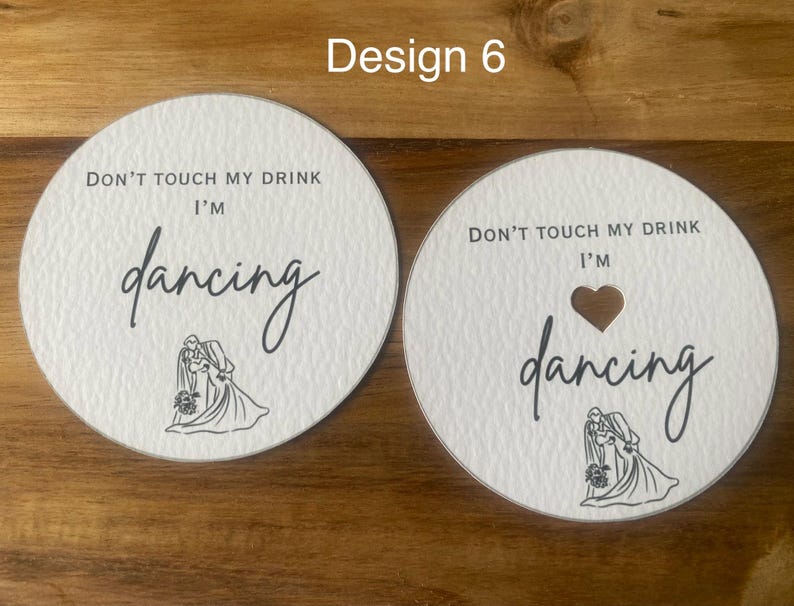 May include: Two white round coasters with the text "DON'T TOUCH MY DRINK I'M dancing" in black script. One coaster has a heart-shaped cutout. Both have a line drawing of a couple dancing.