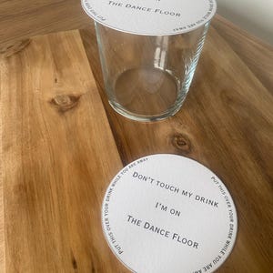 May include: A clear glass tumbler with a white paper coaster on top and another coaster beside it. The coasters have the text "DON'T TOUCH MY DRINK I'M ON THE DANCE FLOOR" printed on them.