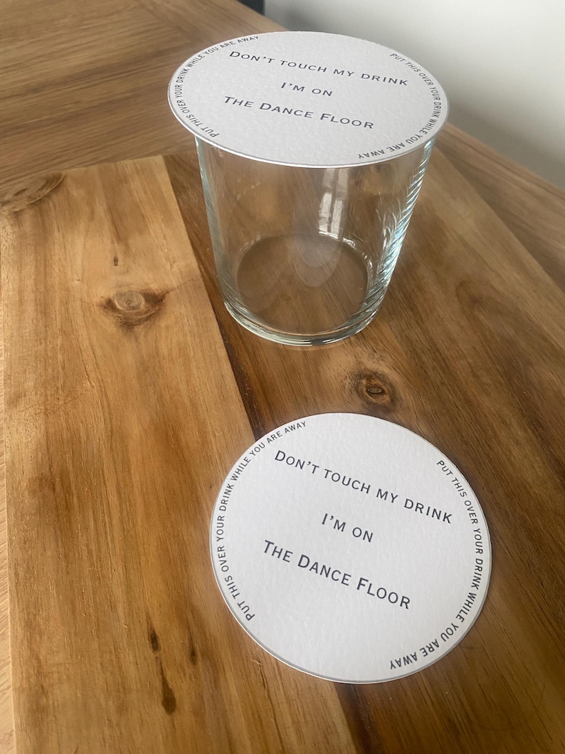 May include: A clear glass tumbler with a white paper coaster on top and another coaster beside it. The coasters have the text "DON'T TOUCH MY DRINK I'M ON THE DANCE FLOOR" printed on them.