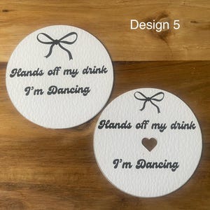May include: Two white, round coasters with black text and bow designs. One coaster has a heart-shaped cutout. The text on both coasters reads "Hands off my drink I'm Dancing".