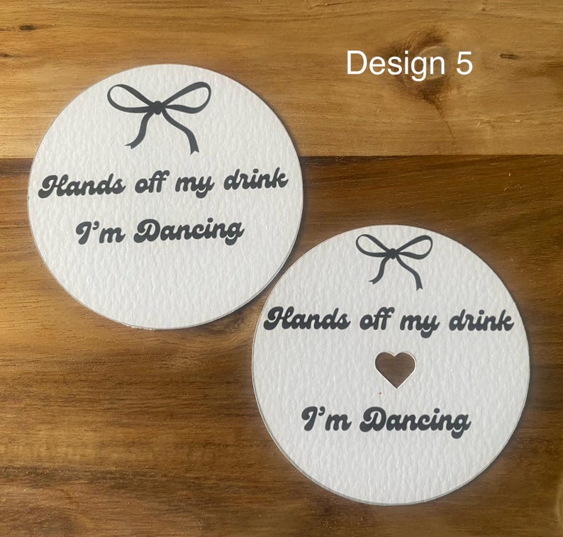 May include: Two white, round coasters with black text and bow designs. One coaster has a heart-shaped cutout. The text on both coasters reads "Hands off my drink I'm Dancing".