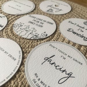 May include: A collection of white, round coasters with black text and illustrations. The coasters feature phrases like "Don't touch my drink, I'm dancing" and "Please leave my drink, I'm on the dance floor." One coaster is personalized with a date.