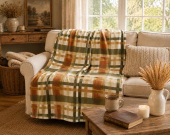 May include: A rectangular throw blanket featuring a plaid pattern in shades of olive green, burnt orange, cream, and tan. The blanket is draped over a light-colored sofa, displaying a soft, smooth texture with a repeating grid of intersecting colored stripes.