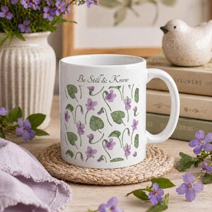 May include: White ceramic mug featuring a violet and leaf pattern, with the text "Be Still & Know". The mug is placed on a round, woven coaster. The mug is a standard size for hot beverages.