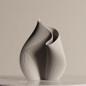 May include: A light gray, abstract sculpture with a flowing, organic form. The piece features vertical, ridged lines that create a textured surface. The sculpture is displayed on a light-colored surface, showcasing its elegant design.