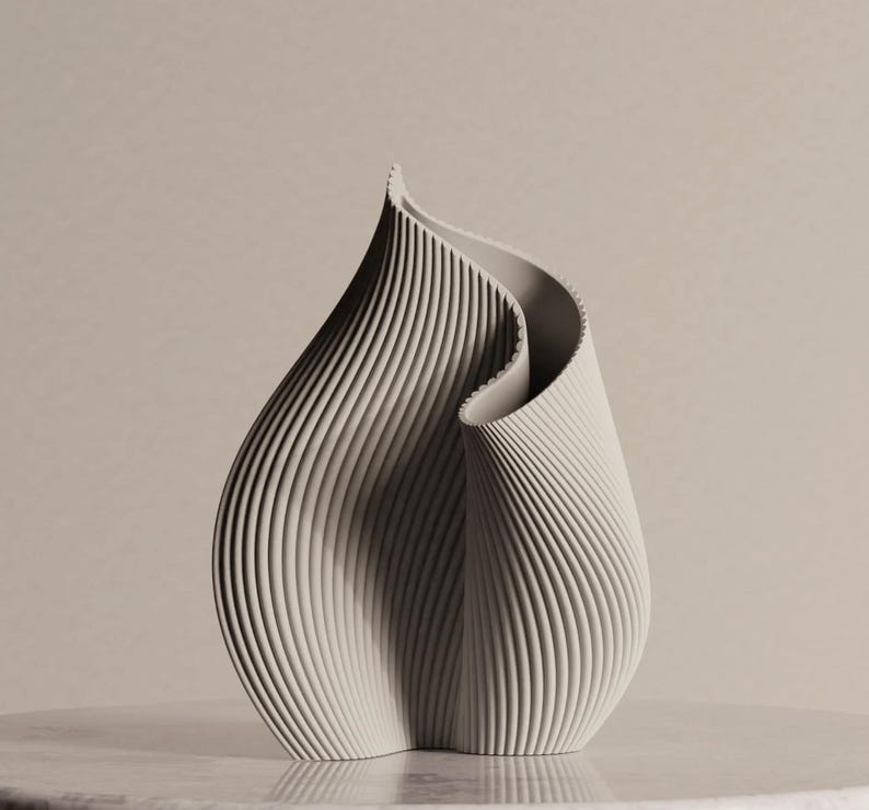 May include: A light gray, abstract sculpture with a flowing, organic form. The piece features vertical, ridged lines that create a textured surface. The sculpture is displayed on a light-colored surface, showcasing its elegant design.