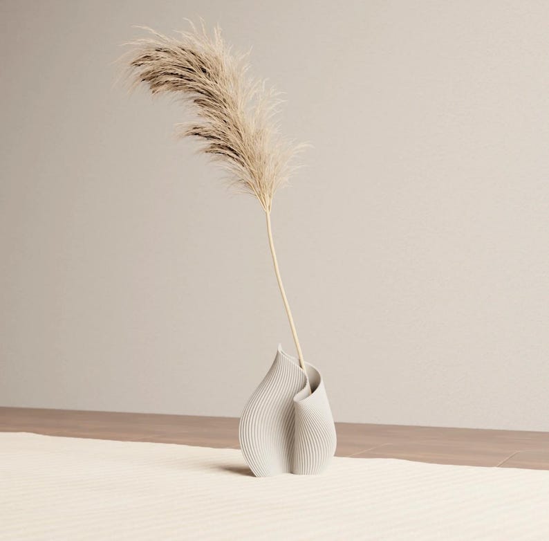 May include: A modern, off-white vase with a unique, curved design, holding a tall, dried ornamental grass. The vase has vertical lines and sits on a cream-colored rug. The background is a neutral beige.