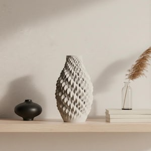May include: A decorative shelf arrangement featuring a black, rounded vase, a textured white vase, a small glass bottle with a dried plant, and a stack of books. The white vase has a unique, layered design. The shelf is made of light-colored wood.