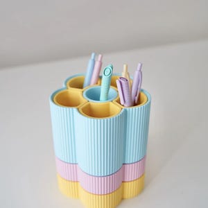 May include: A pastel-colored desk organizer with a flower-like design. The organizer has six compartments in shades of blue, pink, and yellow, holding various pens. The exterior features vertical ribbed detailing. A stylish and functional desk accessory.