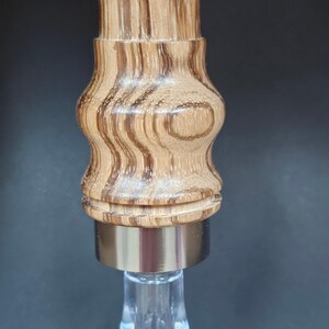May include: A decorative bottle stopper with a wooden top and a clear, faceted glass base. The wooden portion has a light brown and dark brown striped pattern. The metal band is silver.
