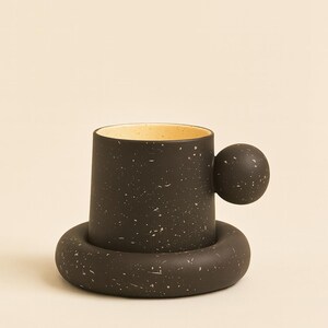 Op de afbeelding: Een zwart gespikkelde keramische mok met een ronde handgreep en een bijpassende onderzetter. De binnenkant van de mok is lichtgeel. Het ontwerp is modern en minimalistisch.