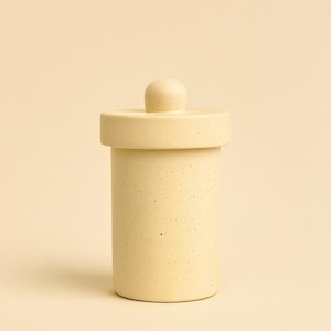 May include: A cylindrical, speckled, light yellow ceramic jar with a matching lid and a small, rounded knob. The jar has a simple, minimalist design, suitable for kitchen or bathroom storage. The background is a solid, light yellow color.