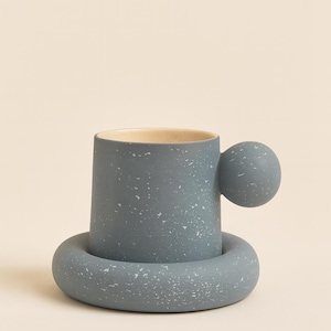 Op de afbeelding: Een gespikkelde blauwe keramische mok met een unieke bolvormige handgreep en een bijpassende schotel. De mok heeft een cilindrische vorm met een lichtbeige interieur. Het ontwerp heeft een gespikkeld patroon, wat een vleugje visuele interesse toevoegt aan dit moderne drinkgerei.
