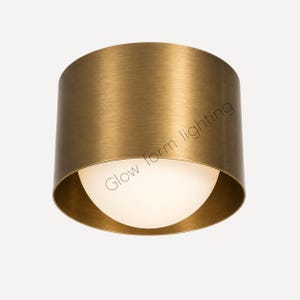 May include: A cylindrical flush mount ceiling light fixture with a brushed brass finish and a central white glass globe diffuser. The text Glow form lighting is printed in a thin, dark font across the side of the metallic outer housing.