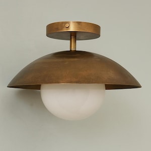 May include: A brass-coloured ceiling light fitting featuring a domed shade and a white globe-shaped light. The fixture has a circular base and a stem connecting the shade and the globe. The design is modern and minimalist.