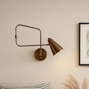 Può includere: Una lampada da parete color bronzo con un paralume a forma di cono. La lampada ha una base circolare e un braccio regolabile. La lampada è montata su una parete di colore chiaro. La lampada è accesa, emettendo un bagliore caldo.
