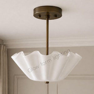 May include: A white, ruffled-edge pendant light fixture with a brass-colored ceiling mount and rod. The fixture has the text "Glow form lighting" on the shade. The light is designed for indoor use.