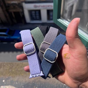 Può includere: Una mano tiene una collezione di cinturini per orologi elastici intrecciati in vari colori: lavanda, verde oliva, nero, grigio e blu navy. Ogni cinturino ha una fibbia color argento e un connettore per orologio abbinato.