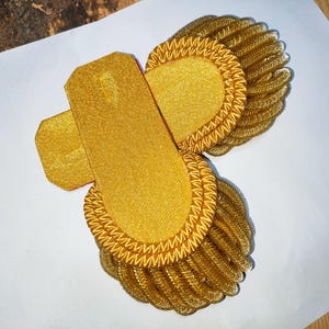 Puede incluir: Charreteras doradas con una parte superior rectangular y una base redondeada y con flecos. La tela brillante y texturizada sugiere que son para un uniforme o vestimenta formal. Se colocan sobre una superficie blanca.