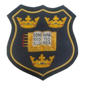 May include: A navy blue embroidered patch in a shield shape, featuring three gold crowns and an open book with the Latin inscription "DOMI MINA NVS TIO ILLU MEA". The patch is outlined in gold thread.