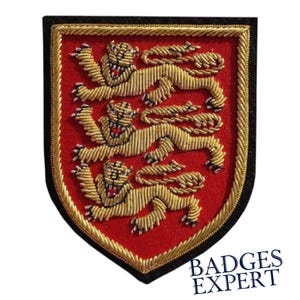 May include: Embroidered patch featuring a red shield with three gold lions passant. The lions are intricately detailed with open mouths and visible teeth. The patch has a gold border and a black edge. The text "BADGES EXPERT" is visible at the bottom.