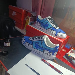 Può includere: Un paio di scarpe da ginnastica Nike Air Force 1 dipinte su misura. Le scarpe presentano una base bianca con dettagli blu e neri in stile cartone animato. Il design include effetti di vernice gocciolante e il logo Nike. Le scarpe da ginnastica sono esposte su una scatola da scarpe Nike rossa.