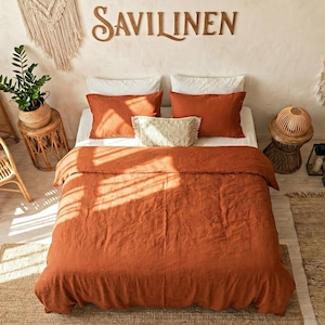 以下が含まれることがあります： 錆色の寝具が特徴の寝室のシーン。掛け布団と枕が含まれています。「SAVILINEN」の文字が壁に表示されています。籐の家具、植物、装飾品が、ボヘミアンスタイルの装飾を完成させています。