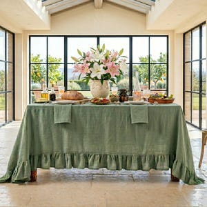 Puede incluir: Una mesa de comedor preparada para una comida, con un mantel de lino verde salvia con borde con volantes. La mesa está puesta con platos, vasos y un jarrón de lirios rosas y blancos. También hay una hogaza de pan y varios platos.