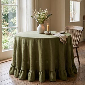 May include: A round, olive green tablecloth with a ruffled edge covers a table. A white vase with flowers and a lit candle sit on top. A wooden chair is positioned to the right of the table, and a window is in the background.