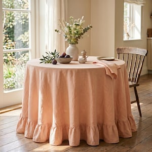 Op de afbeelding: Een rond, perzikkleurig linnen tafelkleed met een geplooide zoom gedrapeerd over een ronde tafel. De tafel is gedekt met een keramische vaas met droogbloemen, een klein schaaltje met verse vijgen, een zoutvaatje en een bijpassend gevouwen servet.