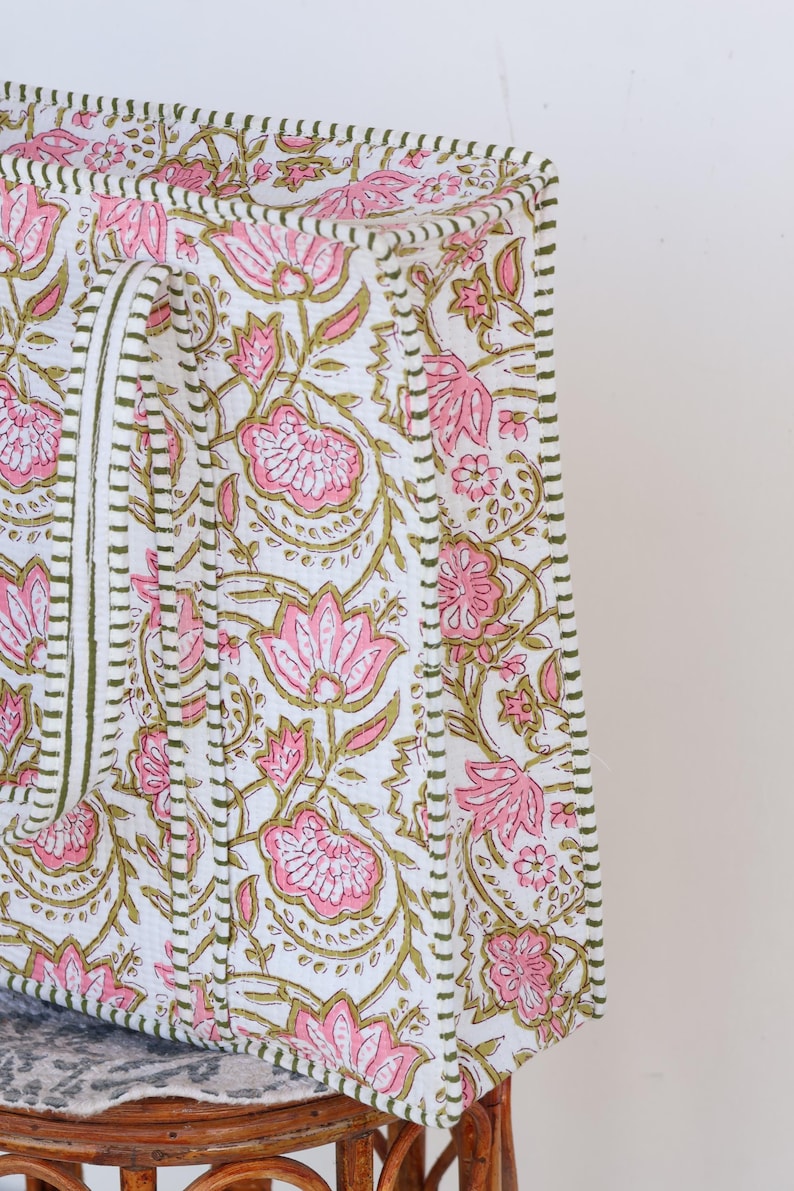 May include: A white tote bag with a pink floral pattern and olive green accents. The bag features a repeating design of stylized flowers and leaves. The edges are trimmed with a green stitched border.