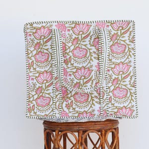 May include: A square tote bag with a floral pattern. The bag features pink lotus flowers and green leaves on a white background. The handles are white with a green and black border. The bag is sitting on a brown wicker stool.