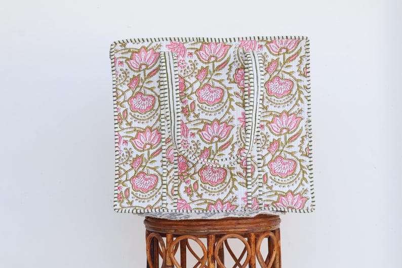 May include: A square tote bag with a floral pattern. The bag features pink lotus flowers and green leaves on a white background. The handles are white with a green and black border. The bag is sitting on a brown wicker stool.
