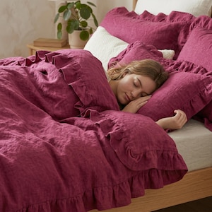 May include: A deep magenta linen duvet cover and matching pillowcases featuring decorative ruffled edges. The bedding set is draped over a bed with a light-colored fitted sheet and a white textured accent pillow, creating a layered appearance.
