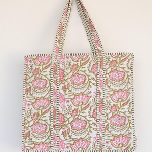 May include: A white tote bag with a pink floral pattern and green accents. The bag features a rectangular shape with two handles and a stitched border. The floral design includes pink flowers and green leaves.