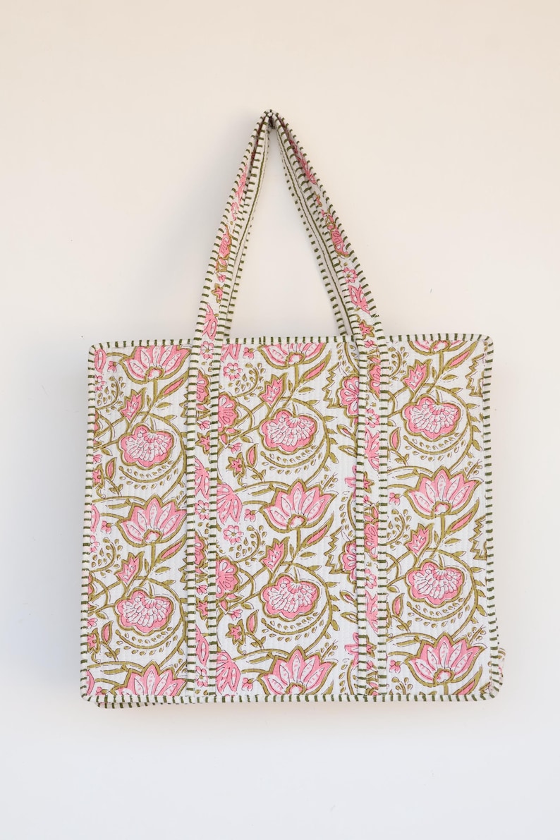 May include: A white tote bag with a pink floral pattern and green accents. The bag features a rectangular shape with two handles and a stitched border. The floral design includes pink flowers and green leaves.