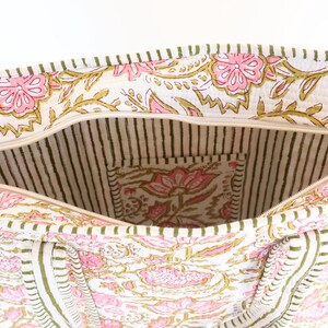 May include: Open tote bag with a floral print in pink and gold on a white background. The interior features a striped pattern and a small pocket with a matching floral design. The bag has a zipper closure.