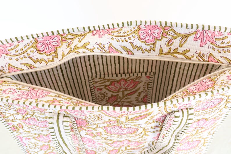 May include: Open tote bag with a floral print in pink and gold on a white background. The interior features a striped pattern and a small pocket with a matching floral design. The bag has a zipper closure.