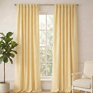 May include: Two pale yellow linen curtains hang on a silver rod in front of a window. A potted plant and a white chair with a cream cushion are on either side of the window. A woven rug covers the floor.